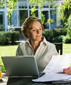 Senior woman sitting in garden with laptop, looking at paperwork