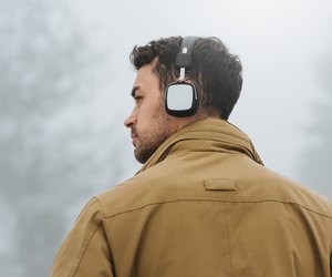 Horizontal portrait of young Caucasian man walking in foggy mountain, listening to music outside. Rear view of Caucasian man wearing trendy coat and bluetooth headphones. People, technology concept.