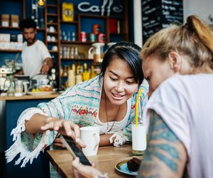 Two Friends Looking Over Pictures On Smartphone In Cafe Together