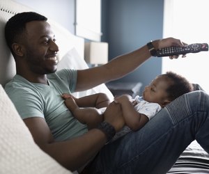 Smiling father with remote control watching TV with baby son in lap on bed