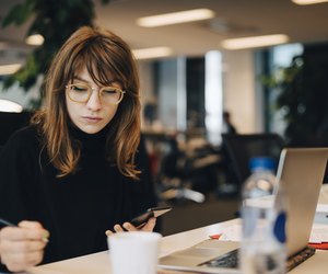 Businesswoman writing while holding mobile phone at desk in office