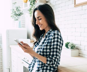 Beautiful cute smiling woman is using smart phone on the kitchen at home.