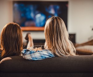 Rear view of two women relaxing on sofa in the living room and watching a movie on TV.