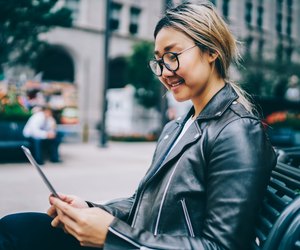 Smiling asian female student resting on bench in downtown reading e-book, positive chinese girl in eyewear using application on tablet for chatting in networks and share multimedia outdoors