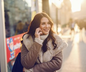 Close up of Caucasian woman with long brown hair dressed in coat using smart phone for call while standing on the street on cold weather. Back light.