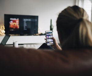 Rear view of teenage girl using smart phone app while watching TV in living room at home