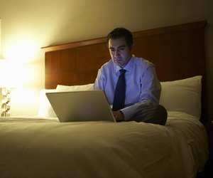 Business man sitting cross-legged on bed in hotel room working on laptop