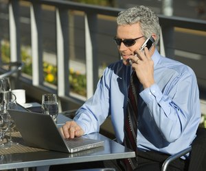 Prime adult Caucasian man in suit sitting at patio table outside looking at laptop and talking on cellphone.