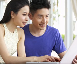 Young Chinese Couple Sitting At Desk And Using Laptop At Home