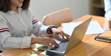 A young secretary is typing on a computer laptop while sitting at the wooden working desk.