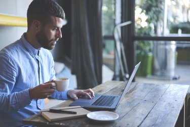 Young skilled male freelancer starting working day early in morning drinking coffee to wake up making new design for website with new advertising content using modern laptop computer and wifi