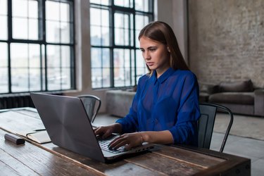 Pretty young brunette working on laptop at wooden table in spacious dark loft studio