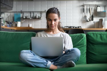 Serious woman using laptop checking news online sitting on sofa