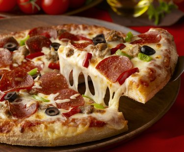 A slice of pizza being lifted from a pizza pie with lots of cheese strings and ingredients in the background.