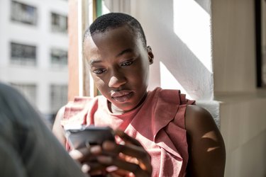 Serious young woman in window sill using cell phone