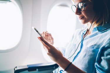 Young female meloman listening favorite songs during flight in first class cabin using mobile playlist and accessory, woman entertaining on airplane board enjoying music in headphones from smartphone