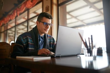 Handsome freelancer businessman working on laptop in cafe. Blogger man updating his profile in social networks with photos sharing with followers multimedia using notebook with wifi. Nerd in glasses