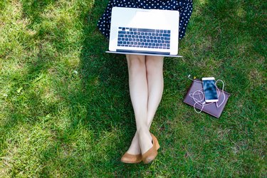 Top view of woman sitting in park on the green grass with laptop, notebook and smartphone, hands on keyboard. Computer screen mockup. Student studying outdoors. Copy space for text