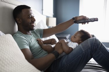 Smiling father with remote control watching TV with baby son in lap on bed