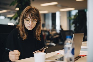 Businesswoman writing while holding mobile phone at desk in office