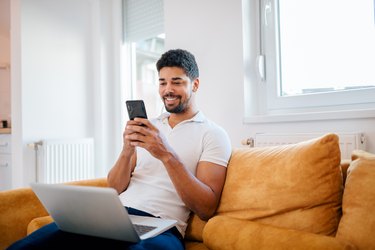 Smiling casual mixed-race freelancer using smart phone and laptop while sitting on the sofa.