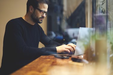 Serious journalist sitting at wooden table using modern devices and searching important information in internet. Student writing article keyboarding text message and concentrated looking at screen