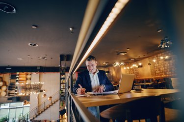 Smiling middle-aged businessman writing down something in agenda while sitting in cafe at the evening. On table coffee and laptop.