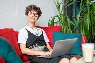 Happy young female in elegant casualwear sitting on couch with laptop in front