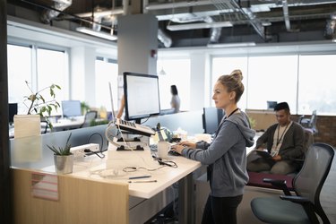 Creative businesswoman standing at desk, using laptop and computer in open plan office