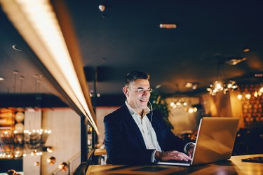 Smiling middle-aged businessman using laptop and having earphones in ears while sitting in cafe at the evening.