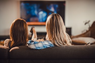 Rear view of two women relaxing on sofa in the living room and watching a movie on TV.