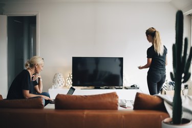 Woman using laptop while teenage daughter standing by television set in living room at home