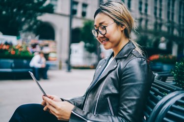 Smiling asian female student resting on bench in downtown reading e-book, positive chinese girl in eyewear using application on tablet for chatting in networks and share multimedia outdoors