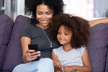 Cheerful mother little daughter spending time at home using smartphone