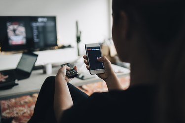 Rear view of teenage girl using phone app and remote control while watching smart TV at home