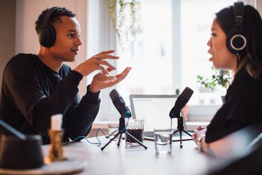 Male blogger gesturing to female wearing headphones sitting at table