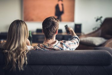 Rear view of a couple relaxing on sofa in the living room and watching a movie on TV.