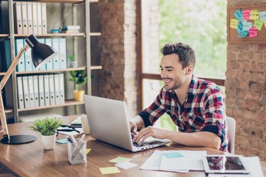 Cheerful young brunet freelancer is smiling, typing on his laptop in nice modern work station at home, in casual smart wear