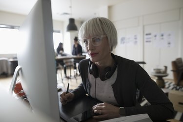 Focused female graphic designer using graphics tablet, leaning toward computer in office