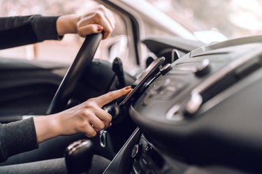 Close up of Caucasian pregnant woman driving car and turning on gps on smart phone. Other hand on steering wheel.