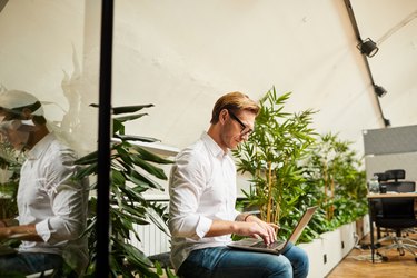 Busy young fashionable male entrepreneur with stubble sitting in open-space office and using laptop while developing online business