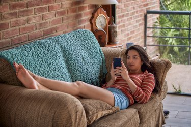 Hispanic young woman laying down on sofa and using smartphone