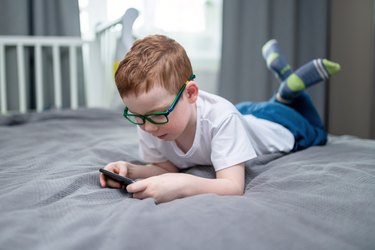 Cute red-haired boy laying down on the bed and playing games on smartphone