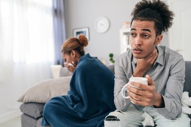 Shot of a sick young african american couple, sitting on the couch at home, the woman is covered with blanket an having a headache, the man holding a cup with hot drink and having a soar throat