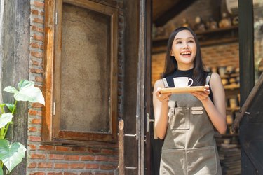 cafe female owner smiling with positive happiness ready to open for customer,asian female woman with apron hand present tray and coffee cup warm welcome condident standing at entrance of coffeeshop