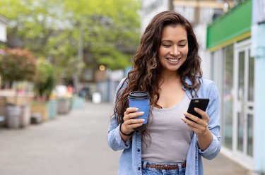 Happy woman drinking a cup of coffee on the move while texting on her phone