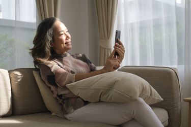 Happy mature woman enjoying browsing social media, using mobile phone on sofa at home.
