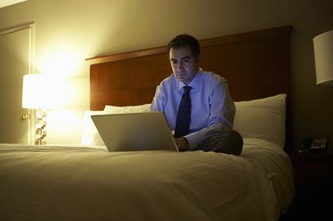 Business man sitting cross-legged on bed in hotel room working on laptop