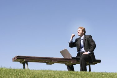 Business man in black suit and blue shirt and tie is holding one arm up in celebration, as he sits on a park bench, next to his laptop