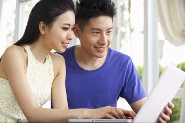 Young Chinese Couple Sitting At Desk And Using Laptop At Home